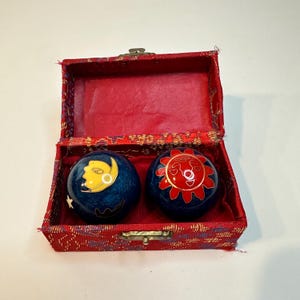 May include: A pair of blue decorative Chinese exercise balls, one with a yellow moon and star design, the other with a red sun design. The balls are inside a red fabric box with a gold clasp.