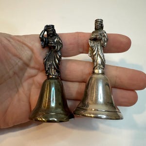 May include: Two antique silver-coloured bells. Each bell features a detailed figure atop a bell-shaped base. One figure is holding their head, the other is standing with hands clasped. The bells are likely made of a metal alloy.