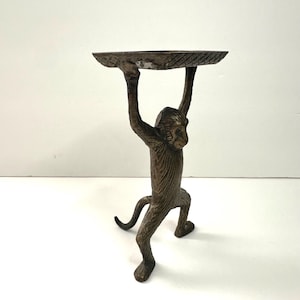 May include: Bronze-colored monkey figurine holding a round, textured tray above its head. The monkey is in a standing position with its arms raised. The tray has a slightly worn appearance. The figurine is on a white surface.