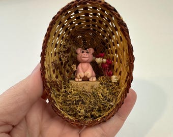 Puede incluir: Una pequeña cesta ovalada de mimbre con una figura de cerdo rosa en su interior. El cerdo está sentado sobre una base de madera, rodeado de plantas secas y pequeñas flores rojas y blancas. La cesta se sostiene en una mano.