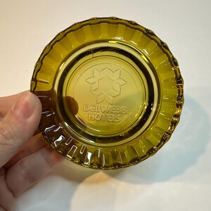 Op de afbeelding: Een vintage olijfgroene glazen asbak met een geschulpte rand. Het midden heeft een bloemmotief en de tekst "Del Webb Hotels". Een klassiek hotelmemorabilia.