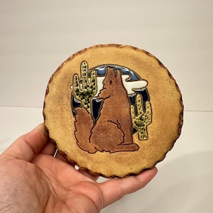 May include: A round, ceramic coaster with a brown coyote silhouette howling at the moon, set against a blue and white night sky. Two green cacti flank the coyote. The coaster has a natural, wood-like edge.