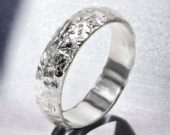 May include: A silver ring with a textured, hammered surface. The ring has a wide band and a shiny, polished interior. The ring's design features an irregular, organic texture, creating a unique and eye-catching appearance. The ring is a piece of jewelry.