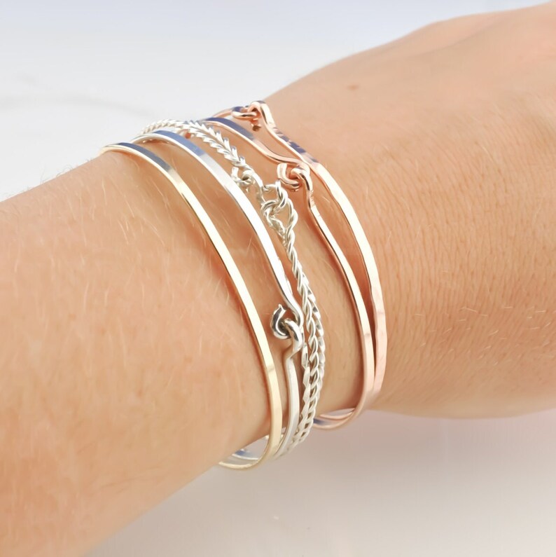 May include: A set of four silver and rose gold bangles stacked on a wrist. The bangles are all different widths and styles, including a twisted rope design and a flat, hammered design.