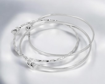 May include: Three silver bangles, one with a twisted design, stacked on a white surface.