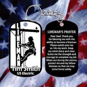 May include: Black and white keychain with a silhouette of a lineman climbing a utility pole. The other side of the keychain has a black background with white text that reads "Lineman's Prayer" and a prayer for linemen. The text on the front of the keychain reads "Lineman Tim Smith US Electric".