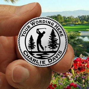 May include: A silver golf ball marker with a black silhouette of a golfer, two pine trees, and the text "YOUR WORDING HERE CHARLIE DAVIS".