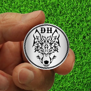 May include: A silver round coin with a black and white wolf head design and the letters "DH" in the center.
