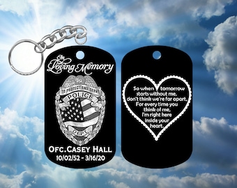 Op de afbeelding: Zwarte metalen sleutelhanger met een zilveren politiebadge en de tekst "In Loving Memory" en "To Protect and Serve". Het badge heeft een Amerikaanse vlag en de tekst "Police Dept". Onder het badge staat de tekst "OFC. CASEY HALL 10/02/52 - 3/16/20". Aan de andere kant van de sleutelhanger bevindt zich een zwart hart met witte tekst die luidt: "So when tomorrow starts without me, don't think we're far apart. For every time you think of me, I'm right here inside your heart."