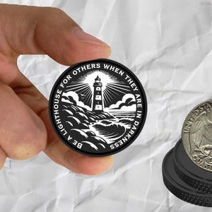 May include: A black round coin with a lighthouse design and the text "BE A LIGHTHOUSE FOR OTHERS WHEN THEY ARE IN DARKNESS." A quarter coin is next to it. The lighthouse is white with black accents.