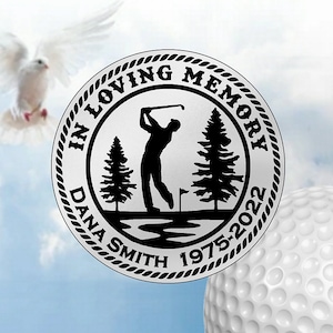 May include: A silver memorial golf ball with a black silhouette of a golfer on a golf course. The text "In Loving Memory Dana Smith 1975-2022" is inscribed around the edge of the ball.