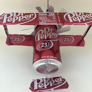 May include: A red aluminum can of Dr Pepper soda, with the words "23 Authentic Blend of 23 Flavors" and "12 fl oz (355 ml)" printed on the can, is transformed into a red and silver model airplane. The airplane has a propeller and wings made from the Dr Pepper can.