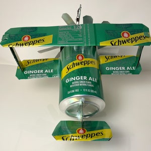 May include: A green and yellow Schweppes Ginger Ale can is transformed into a model airplane. The wings and tail are made from the top and bottom of the can. The can is labeled "Schweppes Ginger Ale" and "Caffeine Free".
