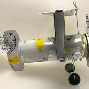 K&ouml;nnte beinhalten: Ein Modellflugzeug aus einer silbernen Dose White Claw Hard Seltzer Lemon. Die Fl&uuml;gel sind aus dem Etikett der Dose und der Propeller aus der Oberseite der Dose gefertigt. Das Flugzeug hat schwarze R&auml;der und einen Draht-Heck.