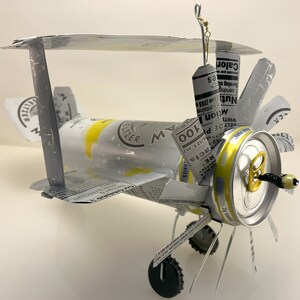 K&ouml;nnte beinhalten: Ein Modellflugzeug aus recycelten Aluminiumdosen. Das Flugzeug ist silberfarben lackiert und tr&auml;gt ein wei&szlig;es, schwarzes und rotes Etikett mit der Aufschrift "RE Facts, 100 Calories, White Claw Hard Seltzer". Das Flugzeug hat einen Propeller aus einem Dosendeckel und eine Angelschnur, die oben befestigt ist.