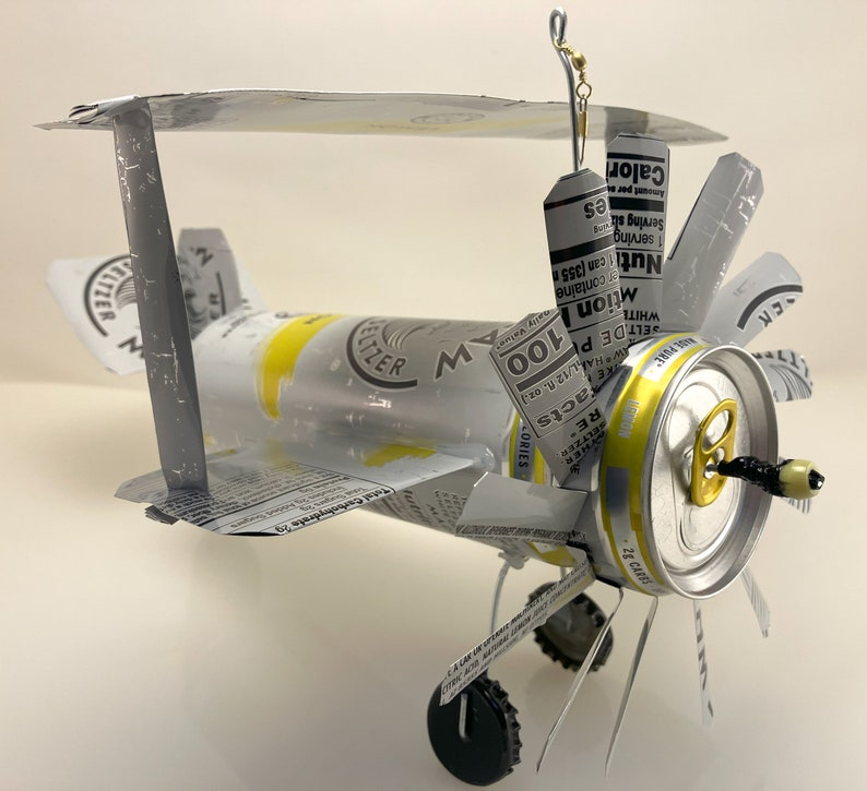 K&ouml;nnte beinhalten: Ein Modellflugzeug aus recycelten Aluminiumdosen. Das Flugzeug ist silberfarben lackiert und tr&auml;gt ein wei&szlig;es, schwarzes und rotes Etikett mit der Aufschrift "RE Facts, 100 Calories, White Claw Hard Seltzer". Das Flugzeug hat einen Propeller aus einem Dosendeckel und eine Angelschnur, die oben befestigt ist.