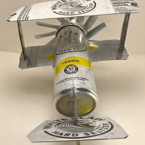 K&ouml;nnte beinhalten: Eine White Claw Hard Seltzer Dose mit Zitronengeschmack, die in ein Modellflugzeug verwandelt wurde. Die Dose ist gelb lackiert und tr&auml;gt ein wei&szlig;es Wellenlogo mit dem Schriftzug "White Claw Hard Seltzer". Die Dose ist mit zwei Fl&uuml;geln aus dem oberen und unteren Teil anderer White Claw Dosen befestigt. Die Fl&uuml;gel sind mit einem Metalldraht an der Dose befestigt.