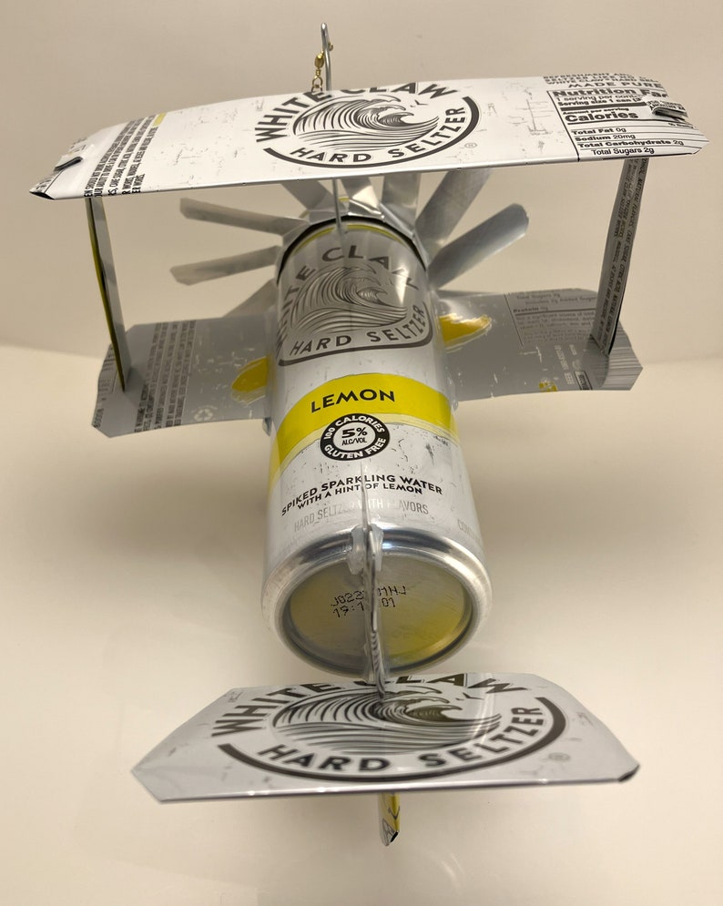 K&ouml;nnte beinhalten: Eine White Claw Hard Seltzer Dose mit Zitronengeschmack, die in ein Modellflugzeug verwandelt wurde. Die Dose ist gelb lackiert und tr&auml;gt ein wei&szlig;es Wellenlogo mit dem Schriftzug "White Claw Hard Seltzer". Die Dose ist mit zwei Fl&uuml;geln aus dem oberen und unteren Teil anderer White Claw Dosen befestigt. Die Fl&uuml;gel sind mit einem Metalldraht an der Dose befestigt.