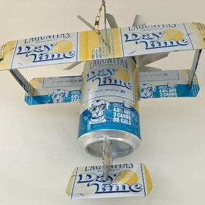 May include: A blue and white can of Lagunitas Daytime IPA beer with a dog illustration and the text "A Crisp Session IPA" and "Only 4.0% ABV 3 Carbs 98 Cals". The can is attached to a cardboard airplane with the text "Lagunitas Daytime" on the wings.