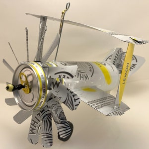 K&ouml;nnte beinhalten: Ein Modellflugzeug aus einer White Claw Hard Seltzer Dose. Das Flugzeug hat einen Propeller aus dem Etikett der Dose und ein Seitenleitwerk aus dem Dosenverschluss. Das Flugzeug ist silberfarben und hat ein gelbes Zitronendesign an der Seite.