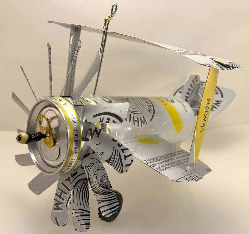 K&ouml;nnte beinhalten: Ein Modellflugzeug aus einer White Claw Hard Seltzer Dose. Das Flugzeug hat einen Propeller aus dem Etikett der Dose und ein Seitenleitwerk aus dem Dosenverschluss. Das Flugzeug ist silberfarben und hat ein gelbes Zitronendesign an der Seite.