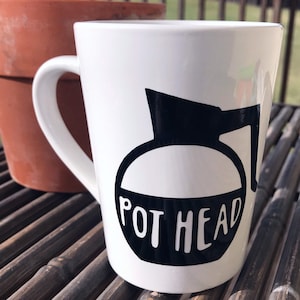 May include: White ceramic coffee mug with a black silhouette of a coffee pot and the text "POT HEAD".