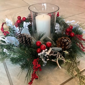 May include: A white pillar candle in a clear glass hurricane sits in the center of a green Christmas wreath. The wreath is decorated with red berries, pine cones, and faux snow.