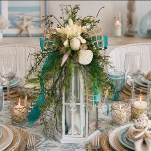 May include: A white metal lantern centerpiece decorated with green foliage, a teal ribbon, and various seashells including a starfish. The lantern is placed on a table set with plates, glassware, and candles, creating a coastal-themed arrangement.