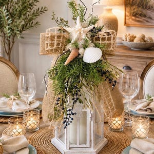 May include: A white wooden lantern centerpiece decorated with a burlap ribbon, a white starfish, a spiral seashell, a white scallop shell, green foliage, and dark blue berries. The lantern contains a large white pillar candle inside.