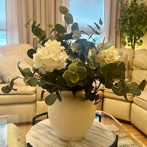 May include: A cream-colored, textured ceramic vase filled with white hydrangeas and eucalyptus greenery. The arrangement sits on a round, distressed wooden table, creating a natural and elegant home decor accent.