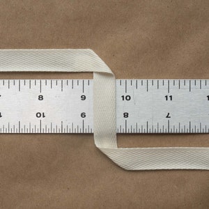 May include: A white cotton twill tape measuring 11 inches long, folded in half and placed over a metal ruler. The ruler is marked with numbers 7 through 12.