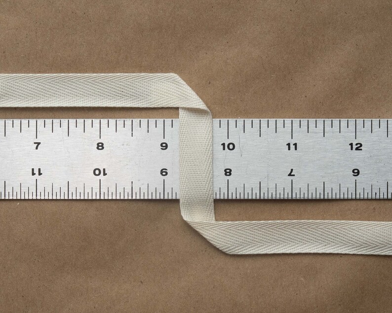May include: A white cotton twill tape measuring 11 inches long, folded in half and placed over a metal ruler. The ruler is marked with numbers 7 through 12.