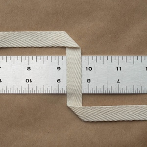 May include: A white cotton twill tape measuring 1 inch wide. The tape is folded in half and placed over a ruler. The ruler shows the tape is 1 inch wide.