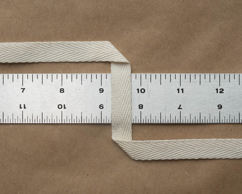 May include: A white cotton twill tape measuring 1 inch wide. The tape is folded in half and placed over a ruler. The ruler shows the tape is 1 inch wide.