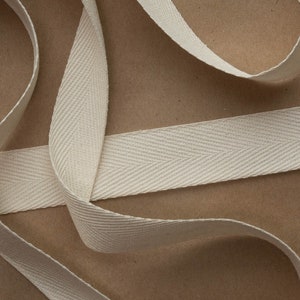 May include: A close-up of a white cotton twill tape with a herringbone pattern. The tape is folded and arranged on a brown surface.