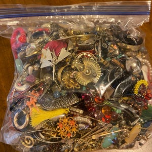 May include: A large clear plastic storage bag filled with a variety of assorted costume jewelry, including gold-tone metal pieces, colorful beads, bright yellow tassels, floral-shaped accents, and various metallic earrings and chains.