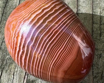 May include: A polished, rounded stone featuring distinct, concentric bands of deep burnt orange, terracotta, and creamy white. The surface is smooth and glossy, highlighting the intricate, wavy, and parallel linear patterns that wrap around the object.