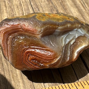 May include: A polished agate stone with layers of red, brown, white, and yellow hues. The natural stone has a smooth, glossy surface and a unique banded pattern. This decorative stone is a natural home decor item.