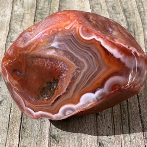 May include: A polished agate stone featuring concentric bands of deep orange, rust red, and white. The center of the stone displays a small, textured cavity filled with tiny, dark, sparkling crystals, showcasing a smooth, rounded exterior surface.