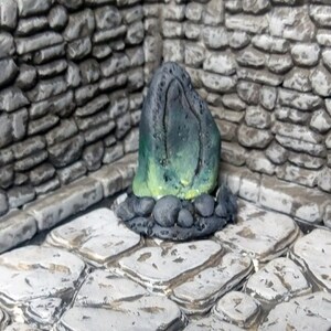 May include: A green and black stone sculpture with a rounded top and a smooth, polished surface. The sculpture is sitting on a base of small, black stones.