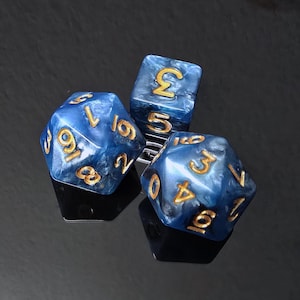 Op de afbeelding: Drie veelzijdige dobbelstenen met een gemarmerde blauwe en witte afwerking en goudkleurige gegraveerde cijfers. De set bevat een twintigzijdige, een twaalfzijdige en een zeszijdige dobbelsteen, allemaal met een glanzende, wervelende textuur op de oppervlakken.