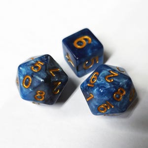 May include: Three blue and gold polyhedral dice. The dice have a marbled appearance with gold numbering. The dice are set against a white background. The dice are used for tabletop games.