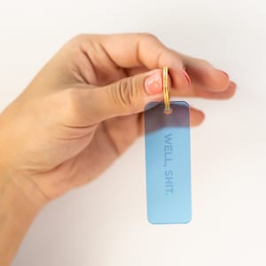 May include: A blue rectangular keyring with the words "WELL. SHIT." in white. The keyring has a gold-coloured ring. The background is white.