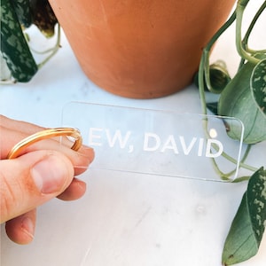 May include: A clear acrylic plant marker with the phrase "EW, DAVID" in white lettering. A gold ring is visible in the image. The background includes a terracotta pot, green plants, and a white surface.