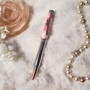 May include: A pen with a pink and white marbled barrel and a silver-colored tip. The pen has a silver rifle charm and a silver bullet-shaped body. A pearl necklace and a pink rose are in the background.