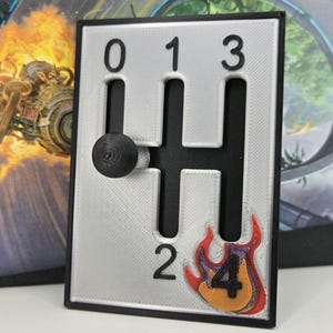 May include: A silver gear shifter with black accents and a red and orange flame graphic with the number "4" inside. The shifter has numbers 0, 1, 2, and 3 on the side.