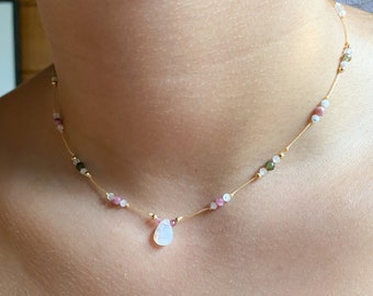 Puede incluir: Un delicado collar de cadena dorada con un colgante de piedra blanca en forma de lágrima. La cadena está adornada con pequeñas cuentas rosas, verdes y blancas.