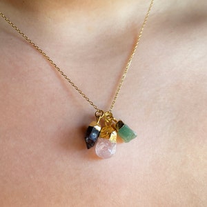Puede incluir: Un collar de cadena dorada con un colgante que presenta tres pequeños cristales en bruto: un verde, un rosa y un negro.