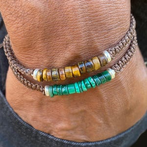 May include: Two macrame bracelets. The top bracelet has brown and yellow beads, the bottom one has green beads. Both bracelets have silver accents and are on a brown cord.