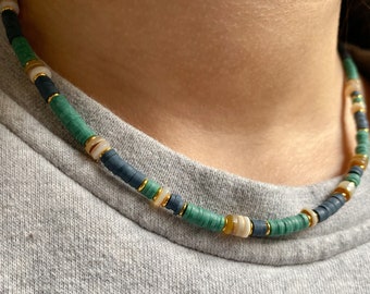 May include: A colorful necklace featuring flat, round beads in shades of teal, navy, and cream, interspersed with gold-colored accents. The necklace is worn against a gray background, highlighting the vibrant colors of the jewelry.